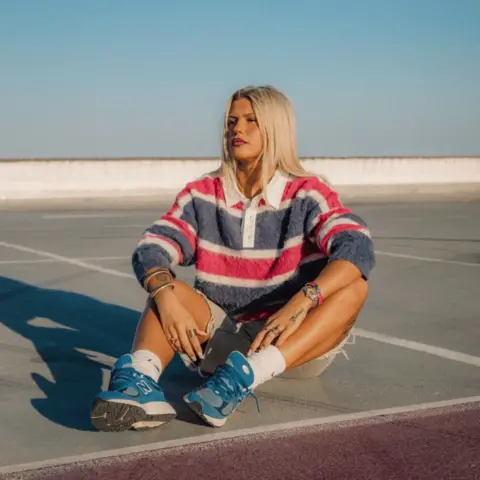 Columbia Records Skye Newman sits on a tennis court, wearing a polo neck sweater with red, white and blue horizontal stripes