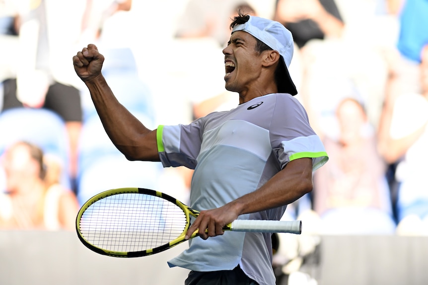 Man in white cap, shirt raises fist while holding tennis racquet