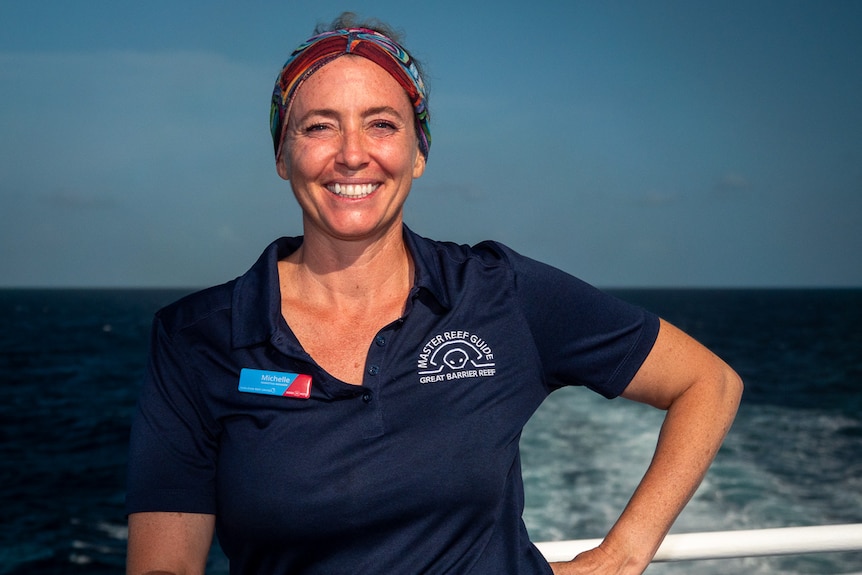 A smiling woman in a headscarf and polo shirt stands on a boat on the ocean.