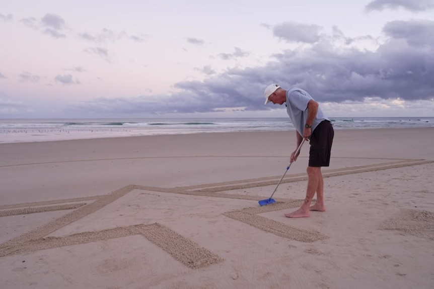 Man holds mop over beach with arrow drawn in sand.