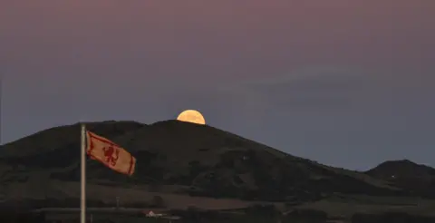BBC Weather Watchers/Louise A moon barely pokes out from behind a dark hill. It is getting dark and the sky is a dark purple/pink. A lion rampant Scotland flag waves on a pole in the foreground