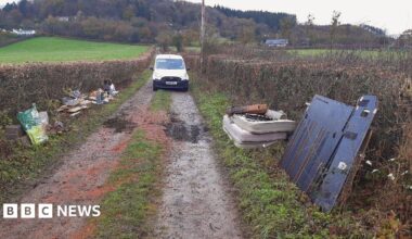A country lane is pictured with mattresses, building waste and metal sheets piled on the grass verges on either side. A white van is parked on the road in shot.