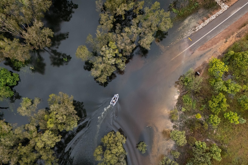 An aerial shot shows waves form in floodwaters as a small boat travels past trees.