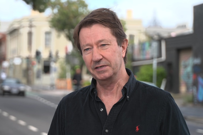 A man with brown hair and a black polo shirt stands on a street.