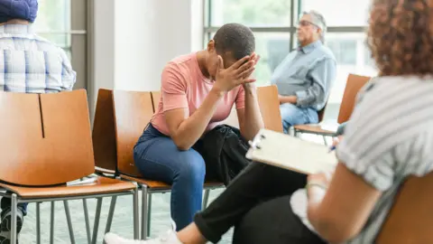 Getty Images A waiting room. One woman sits with her head in her hands, while a man rests his head back on his seat. In the foreground there is a woman writing notes on a clipboard.