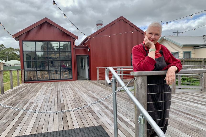A photo of a woman with grey hair standing on a wooden decking and leaning on a fence, with a red building behind her