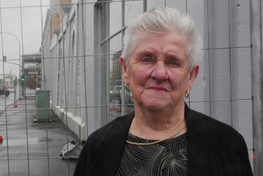 A grey-haired older woman stands in front of a fence with a grey-blue wall with a significant warp behind it.