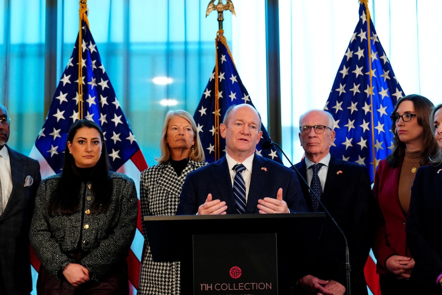  Chris Coons speaks at a lectern with four people behind him and american flags in the background. 