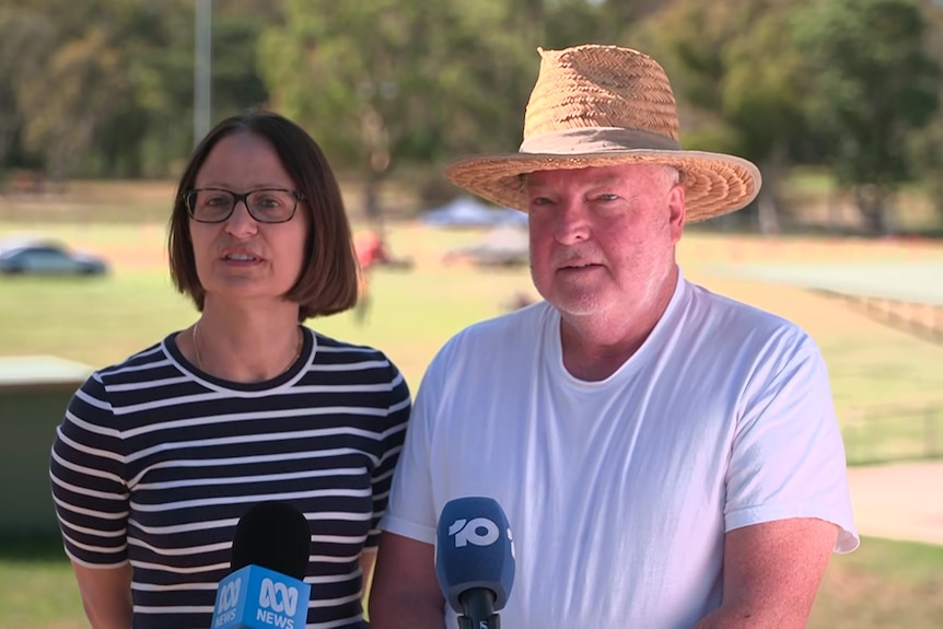 A woman in a black shirt with white stripes and a man in a white tshirt and straw hat stand behind microphones at a park.