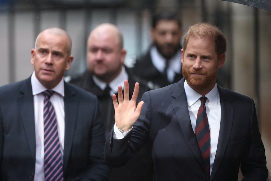 Prince Harry waves in a suit and tie as he walks outside on a London street, tailed by other men