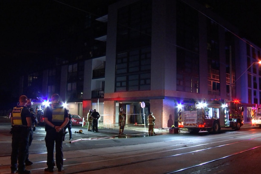 Police and firefighters stand near fire trucks parked on a road beside a multi-storey building at night.