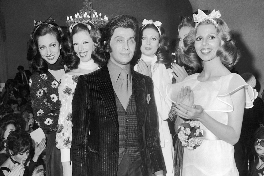 A black and white photograph of a younger Valentino wearing a suit, being clapped by models.