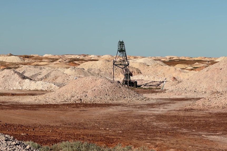 A headframe stands in red dirt with piles of lighter cream dirt around it, against a blue sky. 