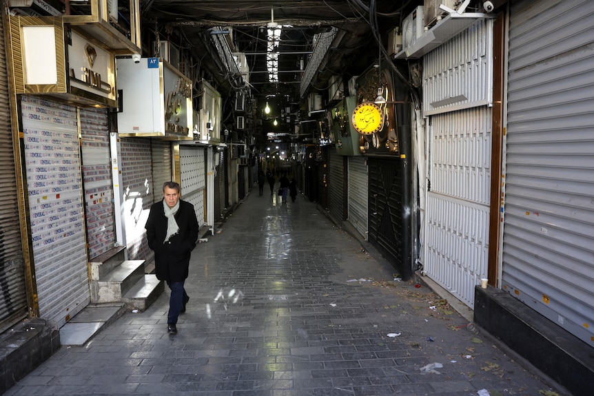 A man in a black coat and grey scarf walks through a deserted Bazaar with lots of closed shops.
