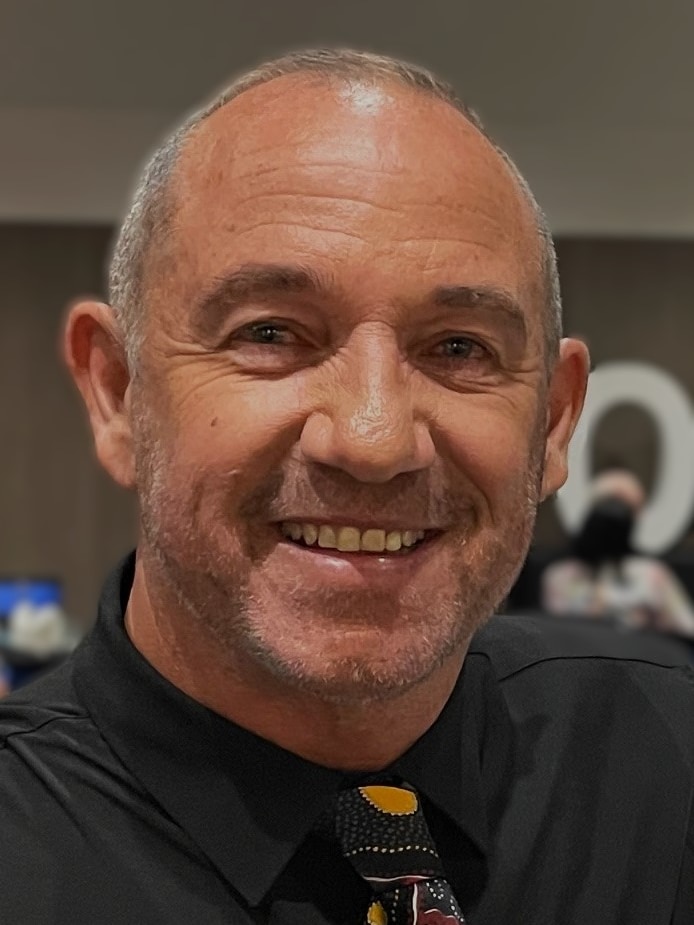 Indigenous man Craig smiling at the camera while wearing a black formal shirt and a tie designed with Aboriginal artwork on it. 