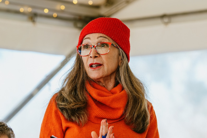 An older woman in an orange jumper and red beanie, with long brown hair and glasses, holding a book and speaking.