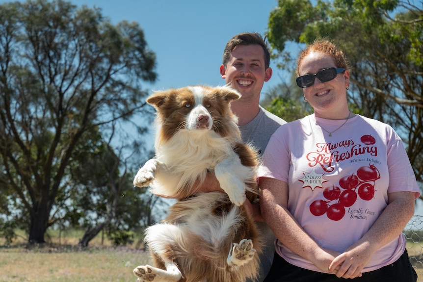 A man holding a dog, next to a woman, with trees in the background