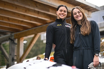 Sarah Blizzard and Desi Johnson, wearing black outfits, smile as they stand next to their sled.
