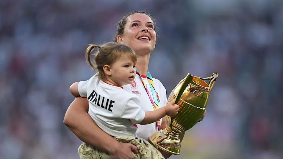 Abbie Ward, wearing her white England shirt, is pictured carrying her daughter Hallie, a toddler who has her hair in pigtails. Both are carrying the gold trophy.
