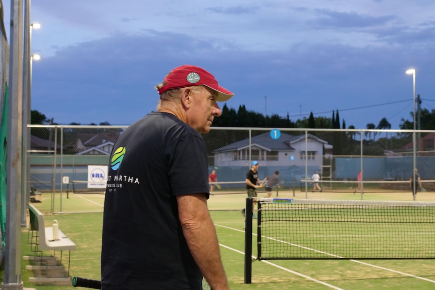 An older man with a black shirt, red cap, watching people play tennis