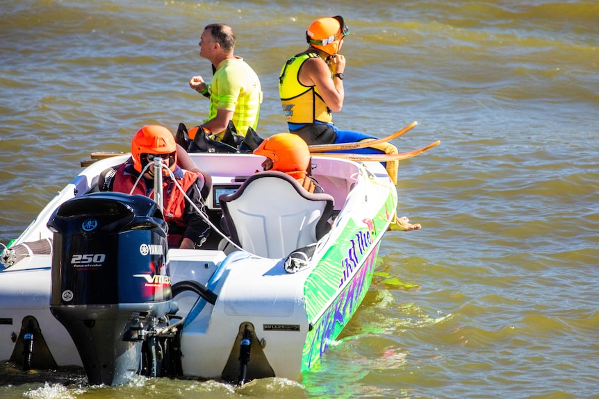 A water skier wearing an orange helmet, prepared to scooch off a boat into a river.