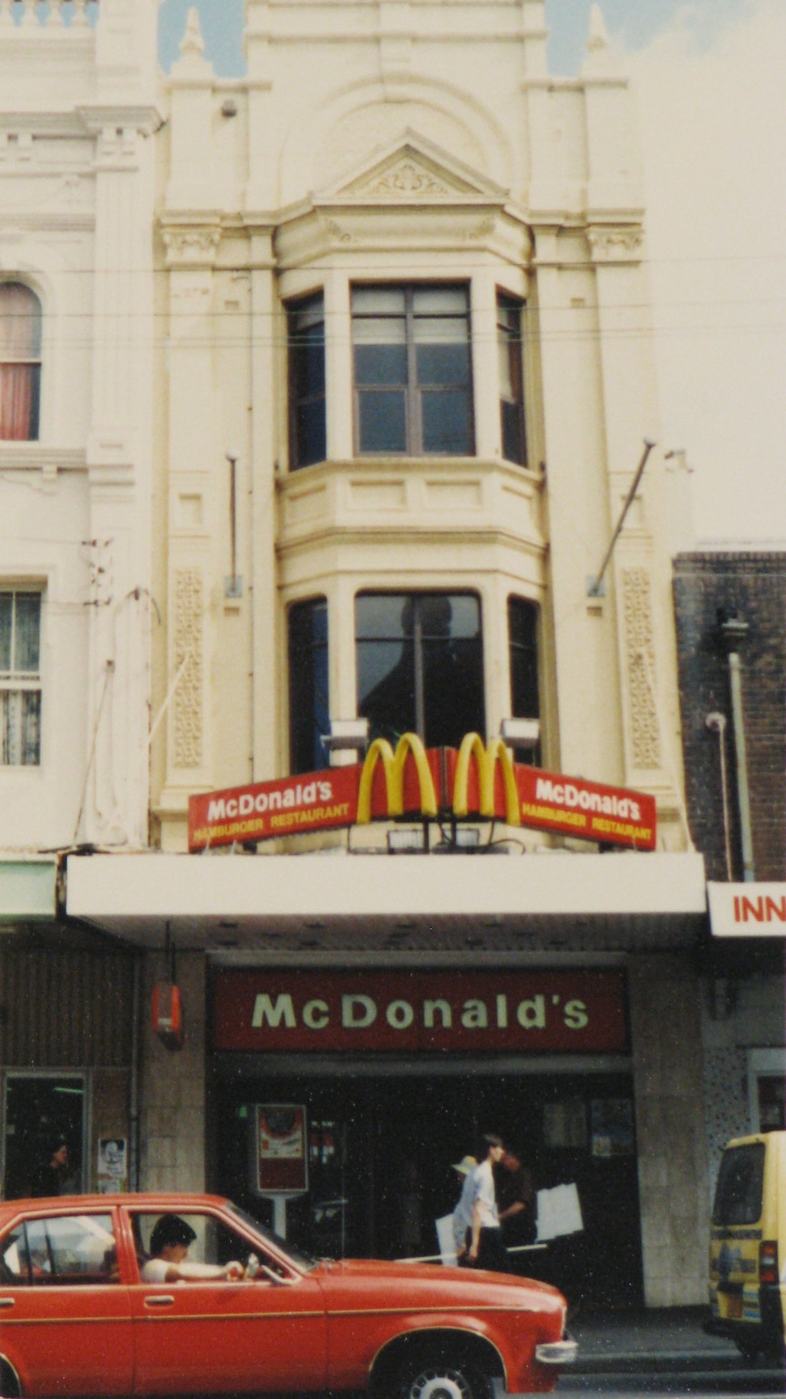 A three-story building with a McDonald's sign under a shopfront verandah, with a red car parked out front.