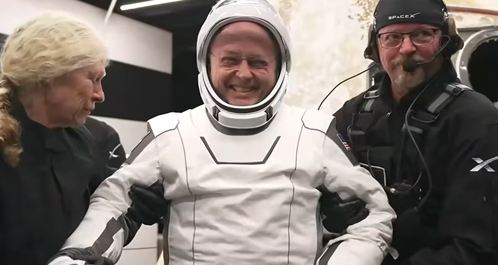 a man in a white spacesuit smiles as he is helped to his feet by two people dressed in black