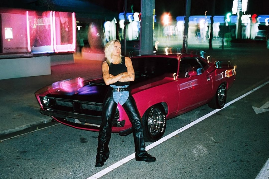 A person stands next to a muscle car with their arms folded.