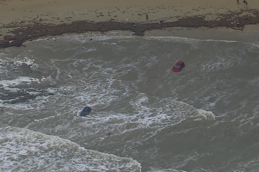 A red and a dark coloured car submerged in water as waves break around them near the sand.