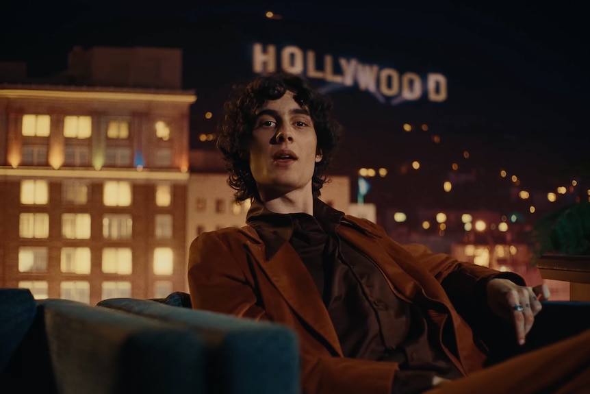 Sombr sits in a velvet suit in a convertible in front of the Hollywood sign at night