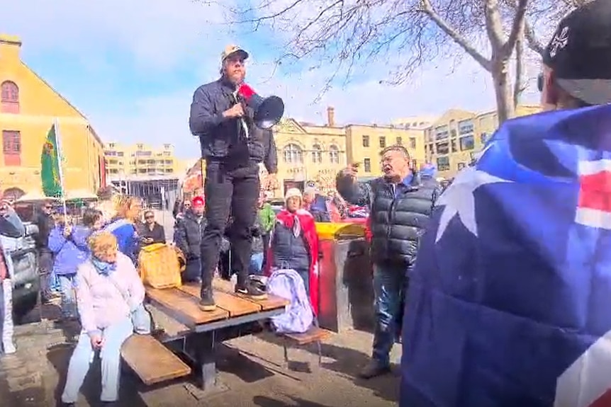 A man holding a megaphone stands on a bench surrounded by people.