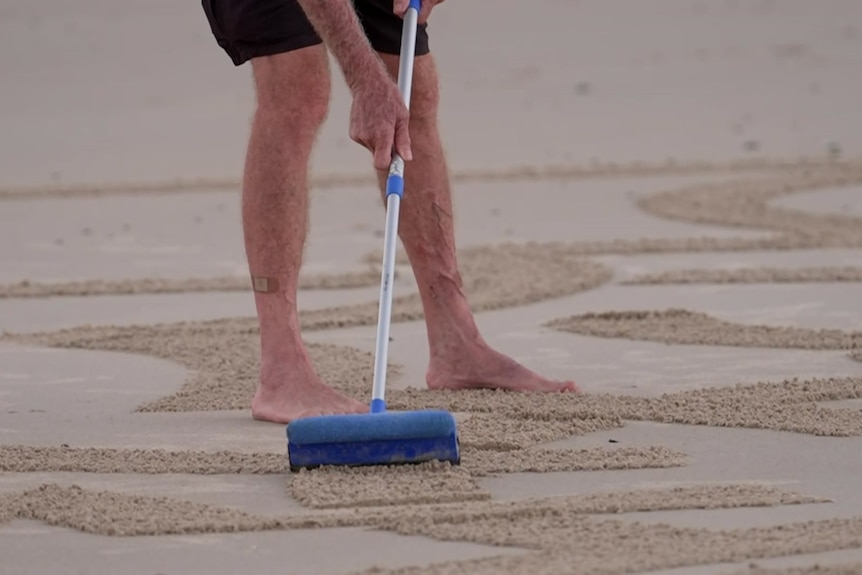 Shot of man's legs and arms as he drags a mop on sand to make patterns.