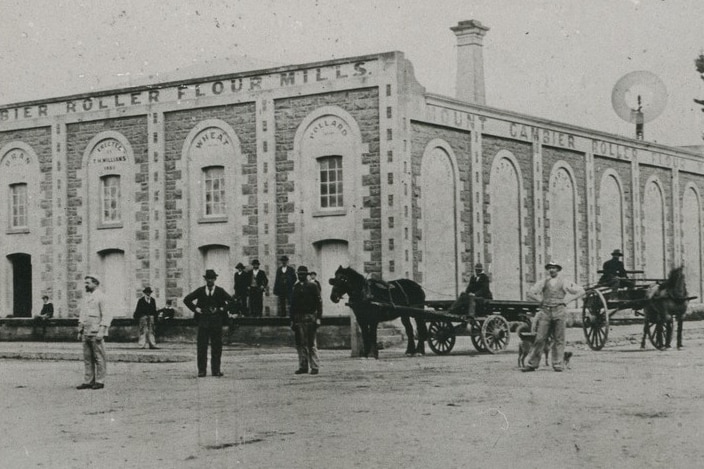 A black and white photo of a brick flour mill, with horse and carts and men standing on the road in front.