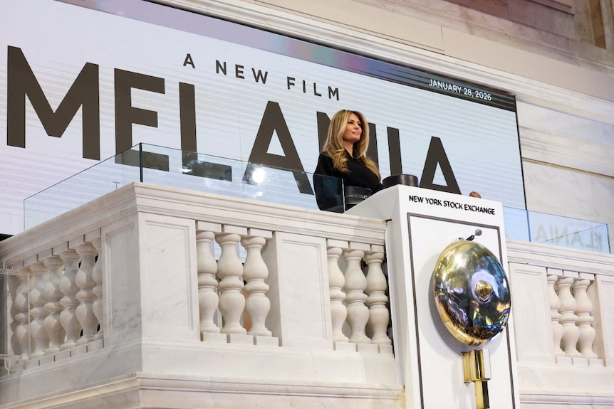 Melania Trump standing on a white balcony at the NY Stock Exchange with a large advertisement behind her for her movie.