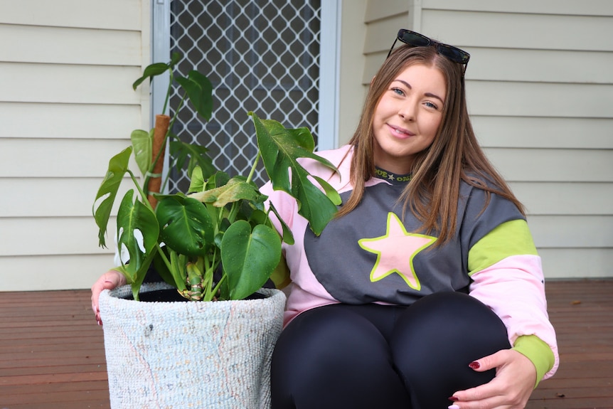 A woman holding a pot of grown monstera plant. The woman is sitting on a wooden porch.