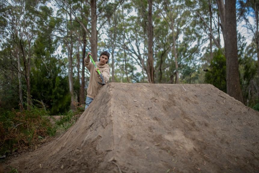 A teenager in a beige hoody and jeans pats down dirt bike jumps with the back of a large shovel in the bush.