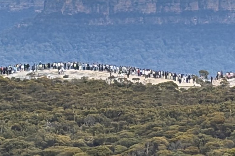 Line of people standing on a white rock, trees and cliffs in the distance.
