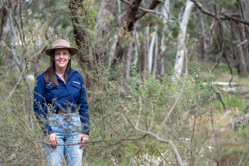 A smiling young woman with long dark hair wears a hat as she stands in a bushy area.