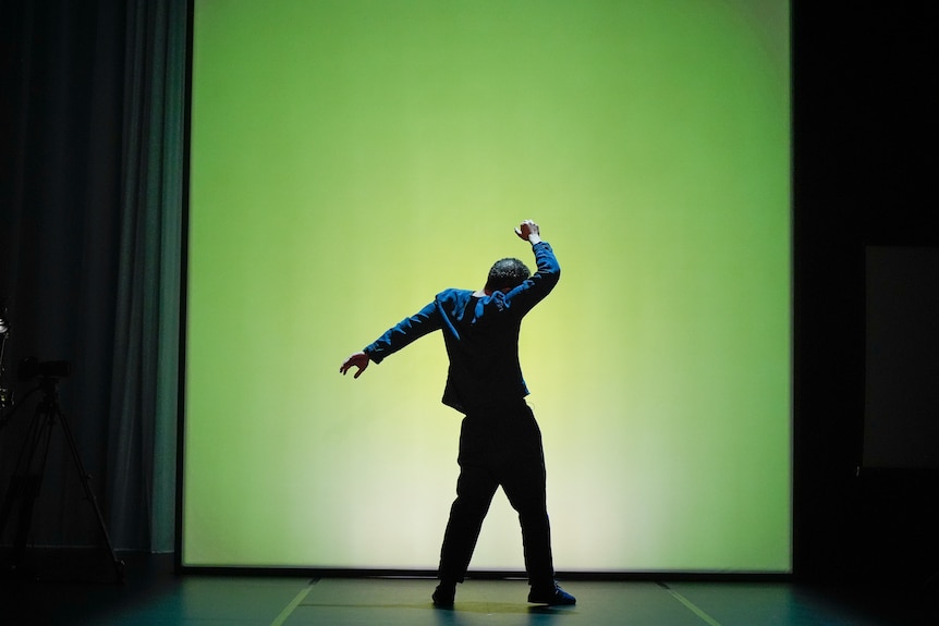 On stage, Khalid Abdalla, an Egyptian British man, 45, dances in front of a green screen, one arm raised above his head.