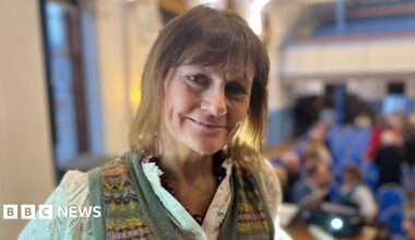 A woman with light brown hair, wearing a white blouse and green tank top, looks at the camera