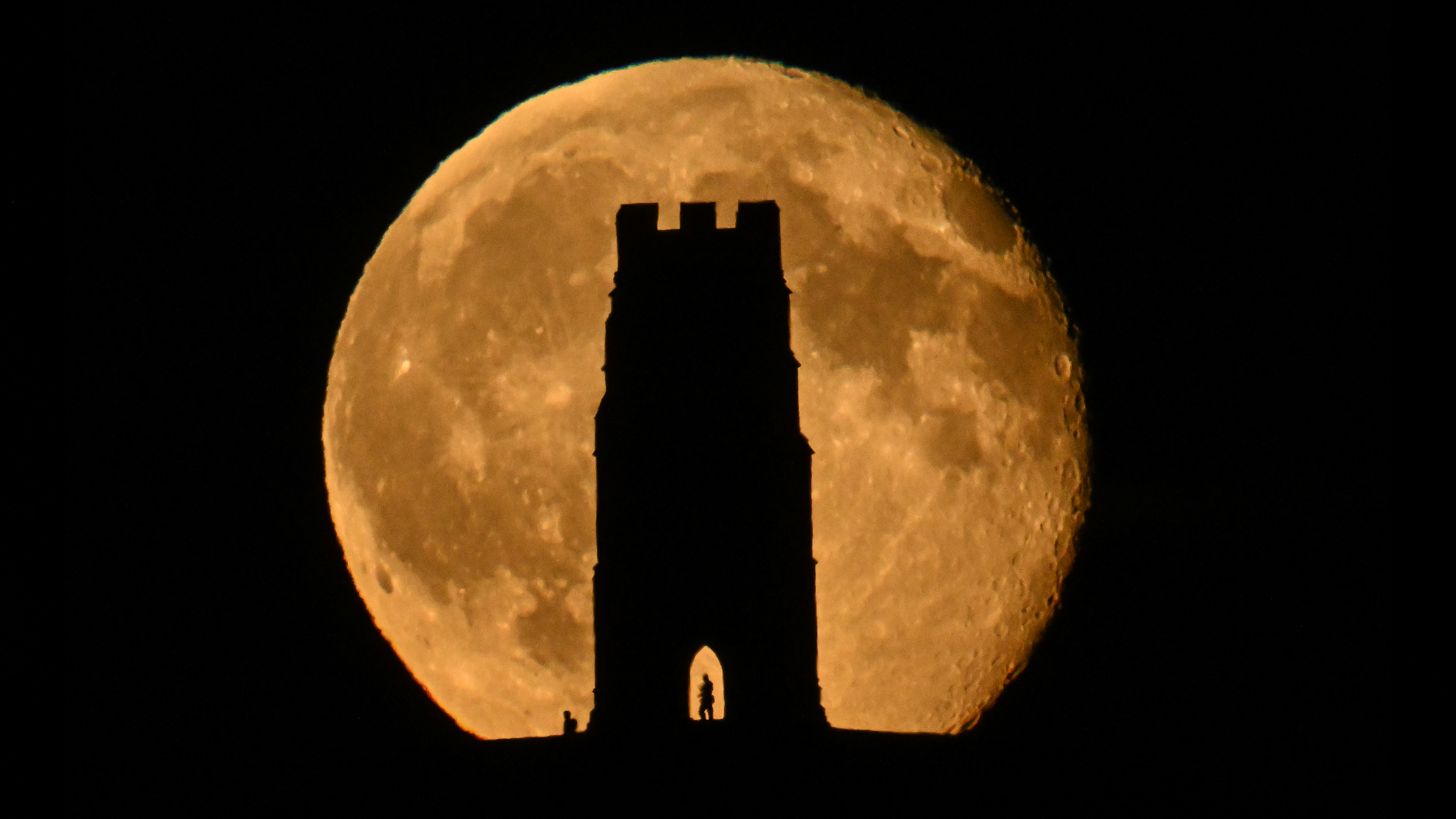 The waning Hunter's Moon is eclipsed by the ancient silhouette of St Michael's Tower atop Glastonbury Tor in Somerset. Distant figures are also in silhouette. The distance of a mile and perspective of an 800mm telephoto lens give this foreshortening effect. No image manipulation, straight shot