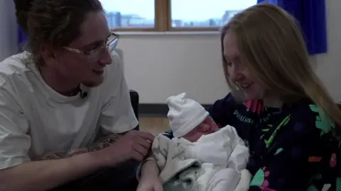 Sonny and Holly sit close together holding a newborn baby wrapped in a blanket.