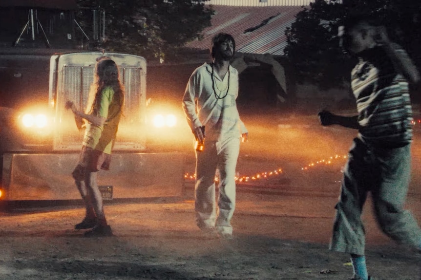 Kevin Parker walks in front of a semi trailer truck in an all white outfit while people dance around him