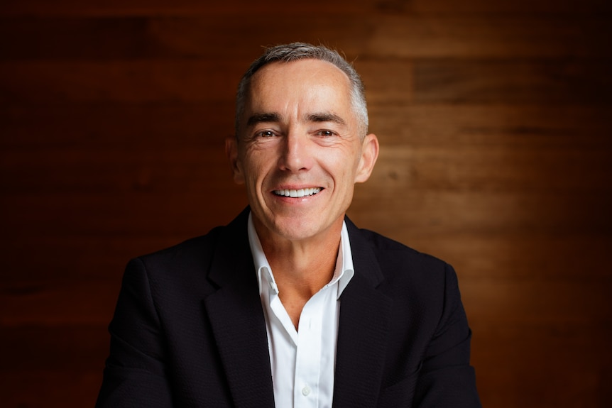 Dean Ormston smiles at the camera wearing a white collared shirt and black jacket.