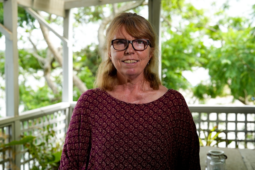 A woman smiles at the camera from outside on a balcony. She wears a maroon jumper and black glasses.