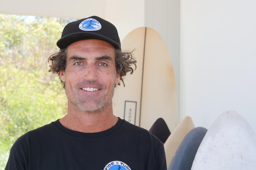 Angus in  black shirt and cap smiling directly at camera with a row of surfboards stacked upright behind him.