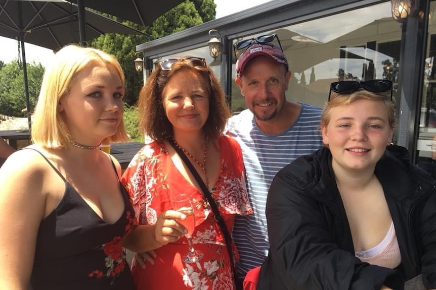Four people standing smiling at the camera. The woman on the far left has blonde hair and winged eyeliner.