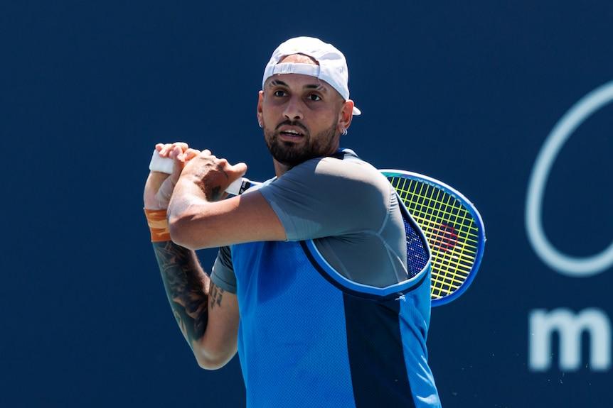 Nick Kyrgios hits a shot, wears a cap backwards, blue tee.