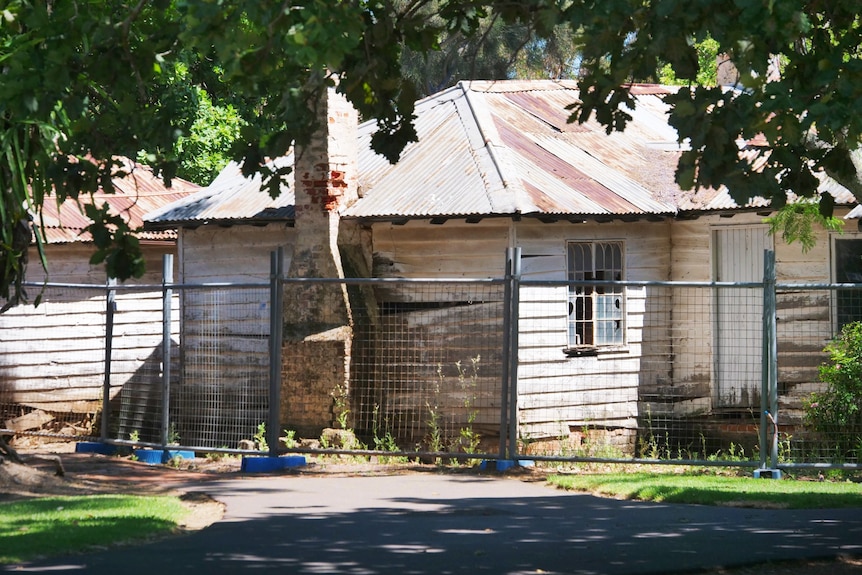 A dilapidated weatherboard cottage with broken window surrounded by fencing