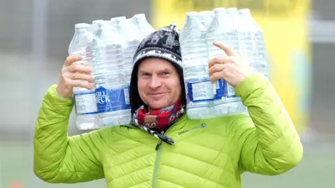 Eddie Mitchell A man wearing a bright lime green jacket, a red patterned scarf and a white, black and grey hat is carrying bottles of water on his shoulders and smiling at the camera